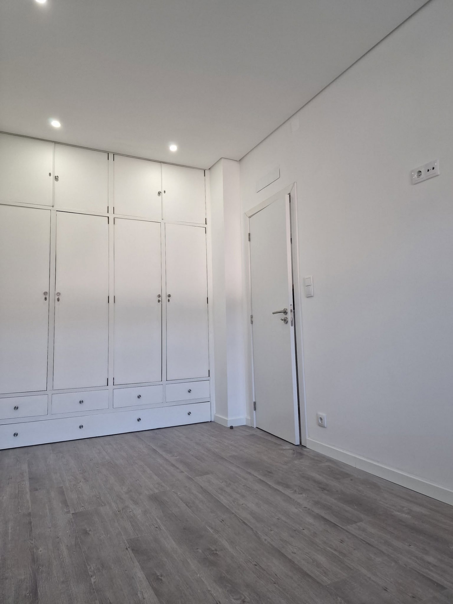 Vista geral do quarto concluído do apartamento T1, com roupeiro embutido branco, pavimento flutuante e teto falso com focos embutidos