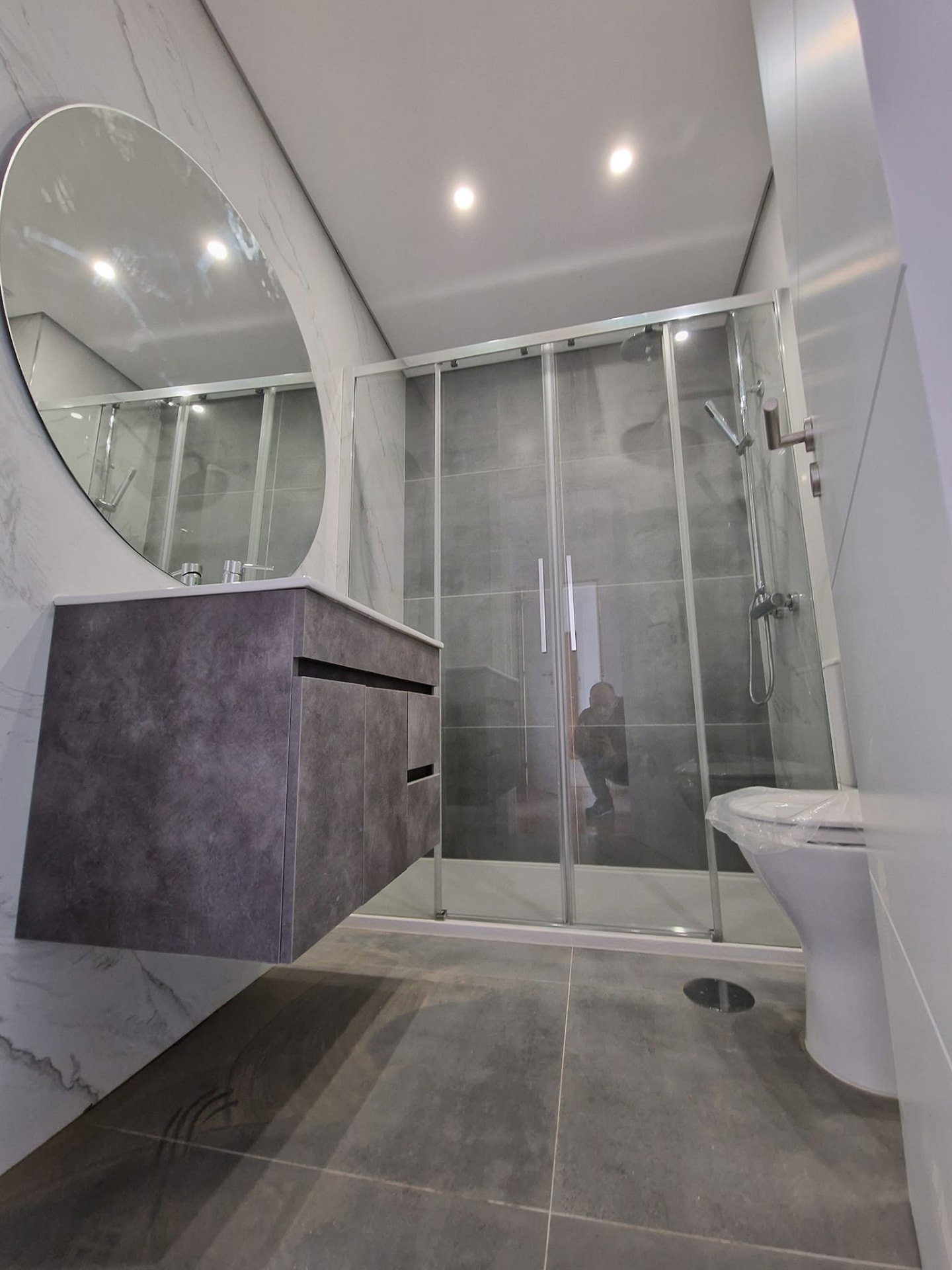 Casa de banho principal do apartamento T3 depois da remodelação, com móvel suspenso, espelho redondo, resguardo de duche em vidro e sanita nova